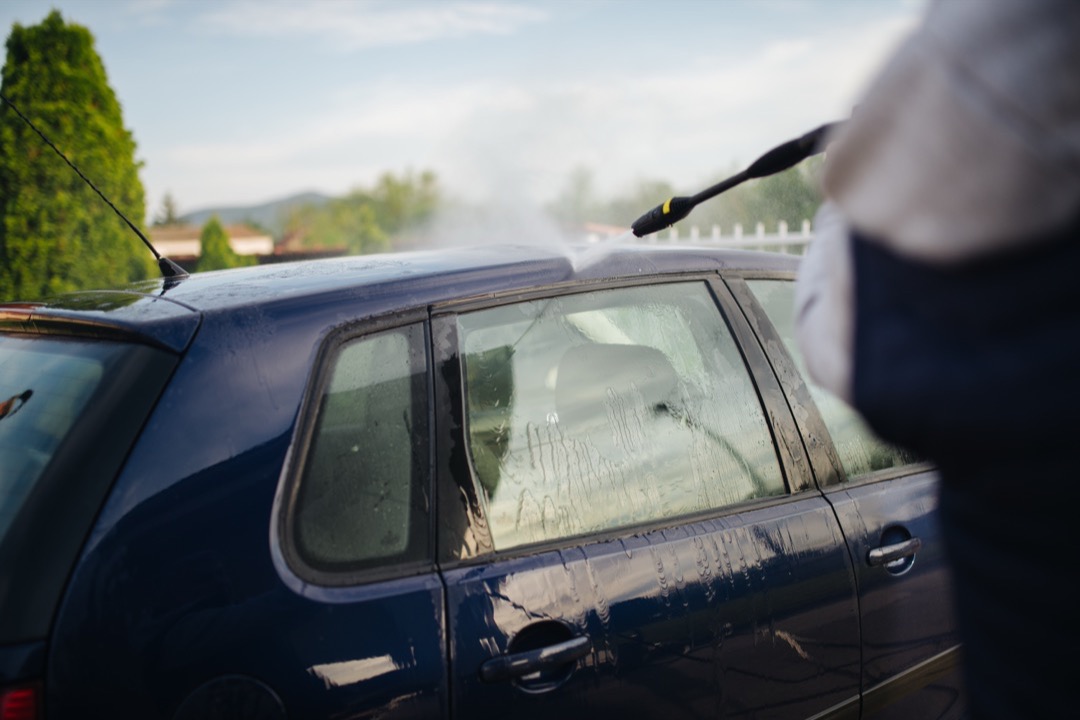 Person washing a silver car with a soapy wash mitt and bucket in a home driveway on a sunny day