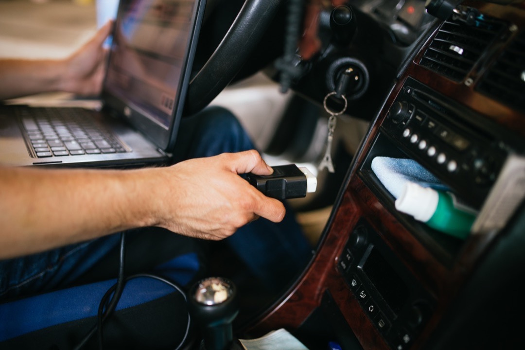 A technician seated in a vehicle cabin holding an OBD2 diagnostic connector with a laptop on his lap, running a scan to diagnose an illuminated check engine light