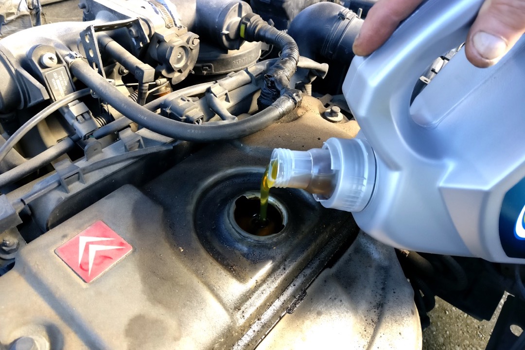 Fresh engine oil being poured from a bottle into a car's oil filler neck during a DIY oil change in the driveway