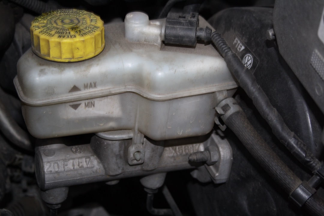 A translucent brake fluid reservoir with a yellow cap and MAX and MIN marks molded into the side, sitting on an aluminum master cylinder in an engine bay