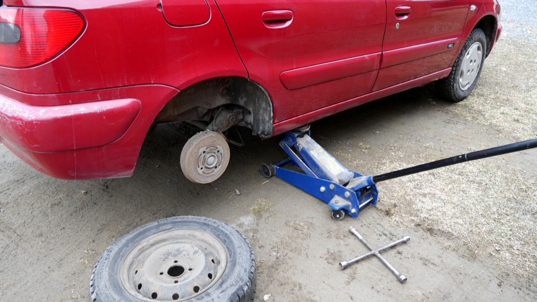 A red hatchback raised on a blue floor jack with the rear wheel removed and the brake drum exposed, the removed wheel and a four-way lug wrench lying on the ground beside it