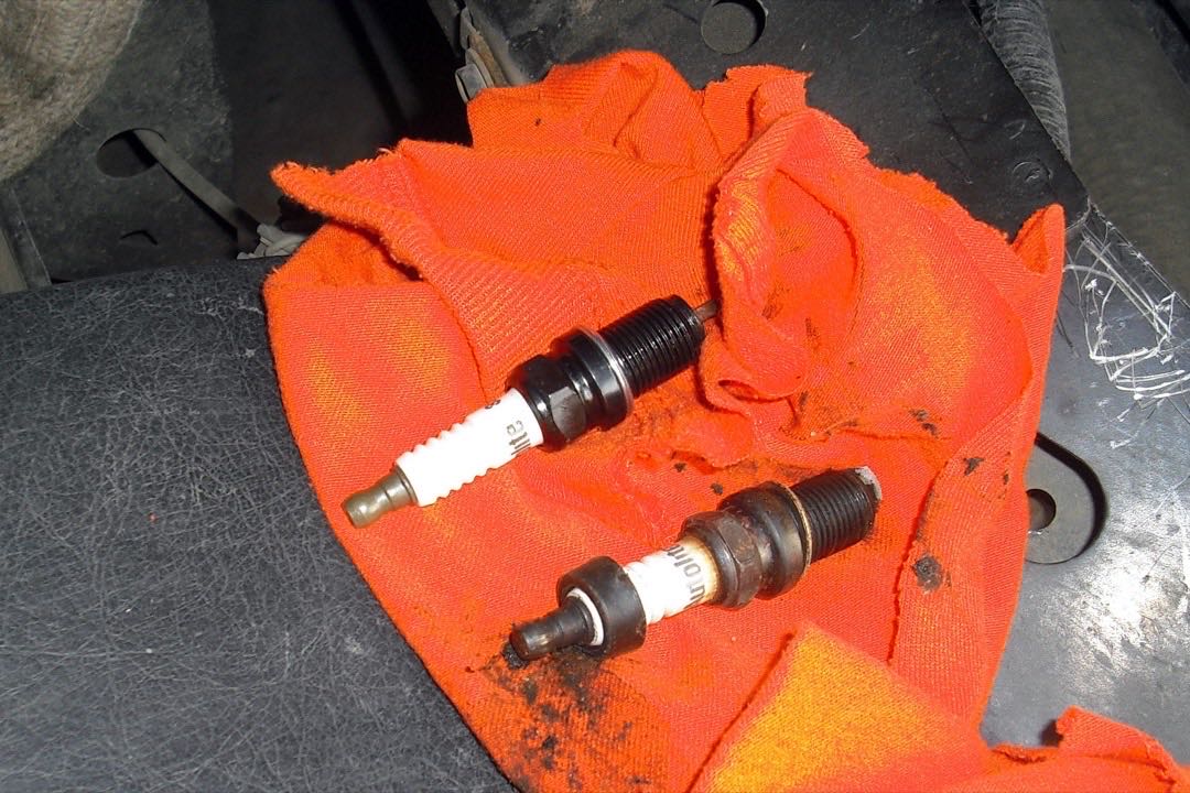 A new spark plug with a clean white insulator lying next to a used, carbon-darkened spark plug on an orange shop rag under the hood of a car