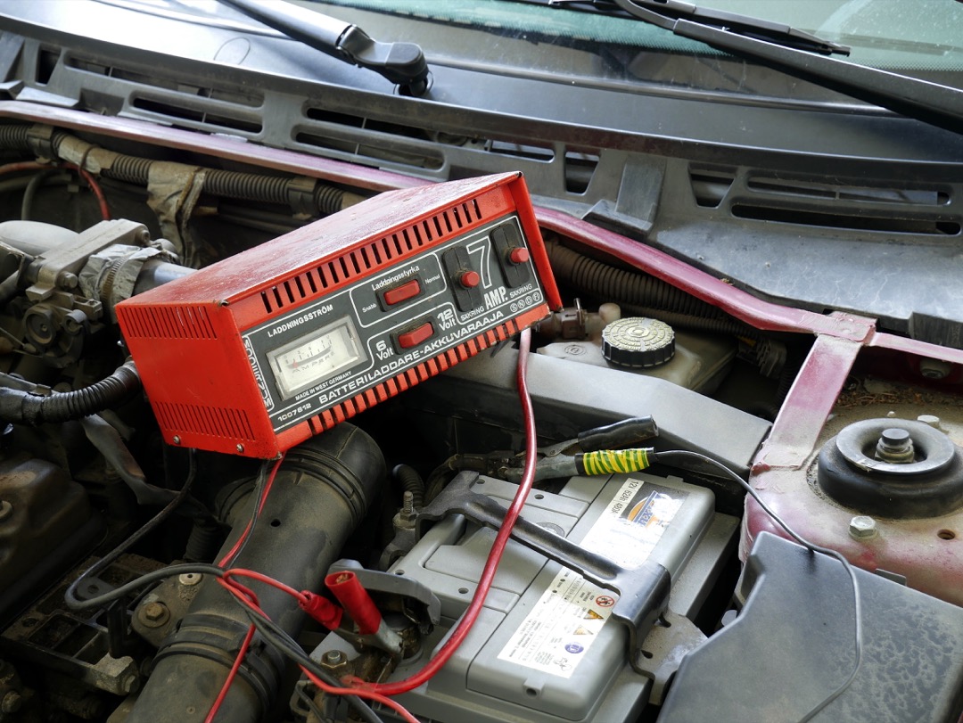 A red analog battery charger with an amp meter sitting in an open engine bay, its red and black clamp leads running down to the vehicle's 12-volt battery