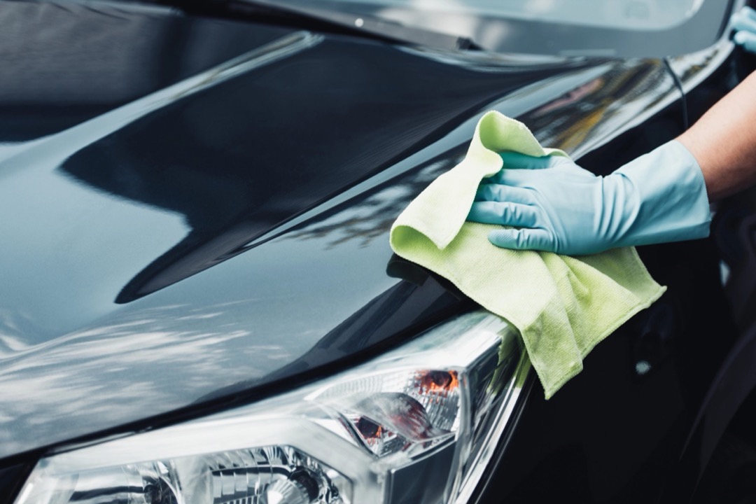 Gloved hand using a green microfiber towel to wipe down the freshly washed hood and headlight of a glossy dark-colored car during a detail
