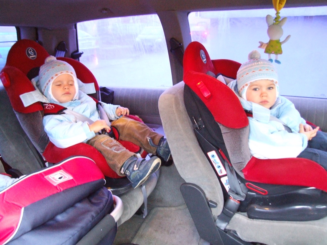 Two young children buckled into rear car seats with the rear side windows visible behind them
