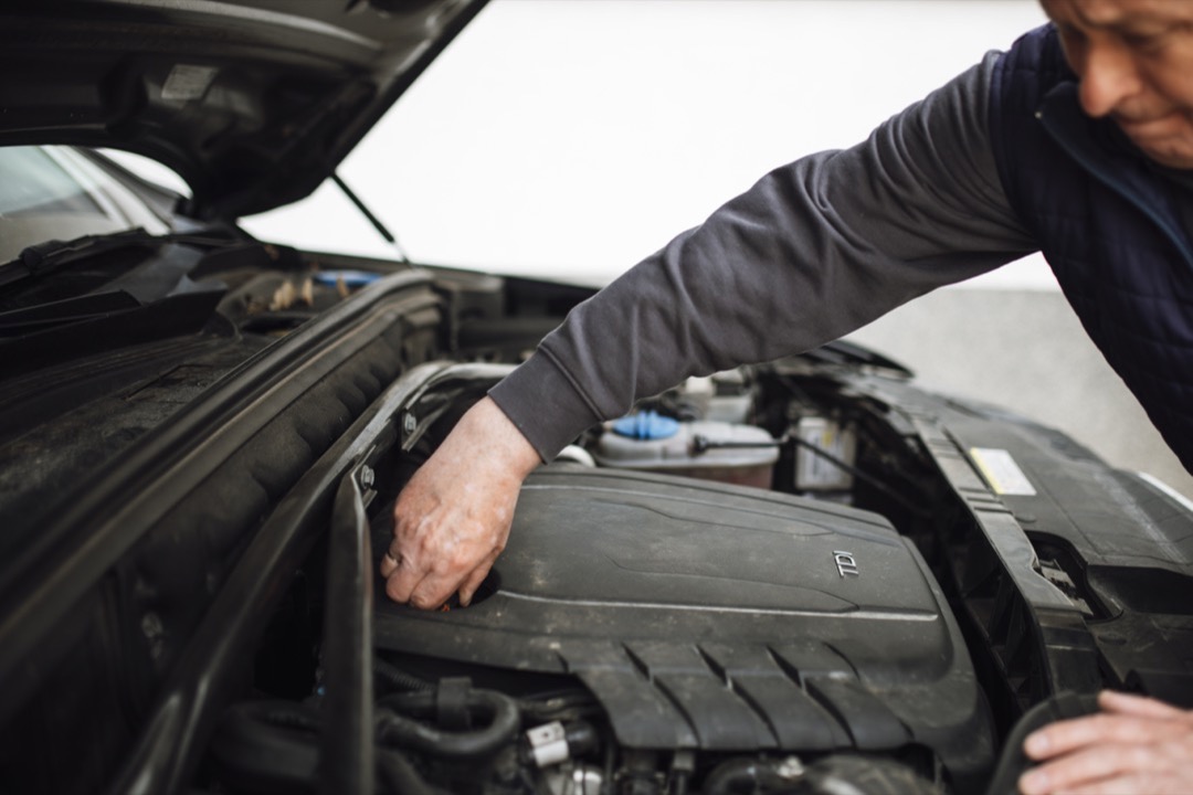A technician working under the open hood of a car, performing a routine maintenance inspection of the engine bay before a trip