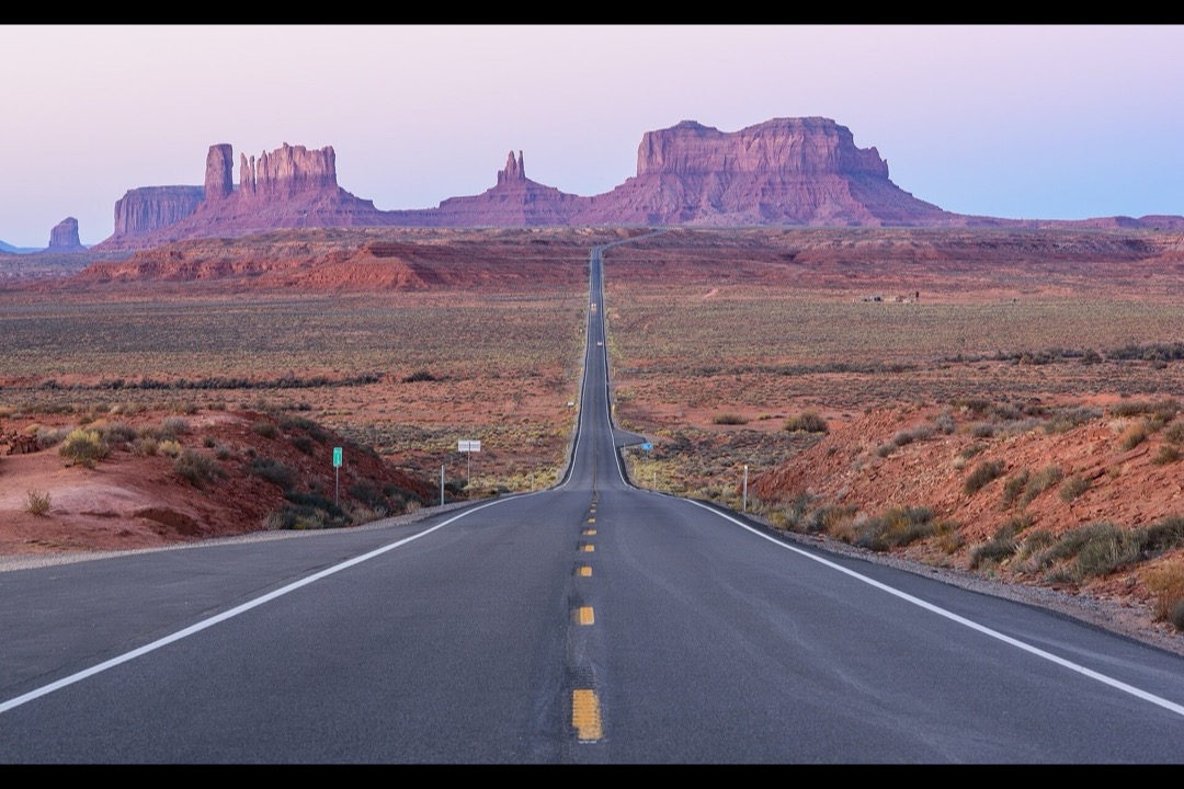A long open highway running straight toward the sandstone buttes of Monument Valley under a clear sky, the classic American road trip view